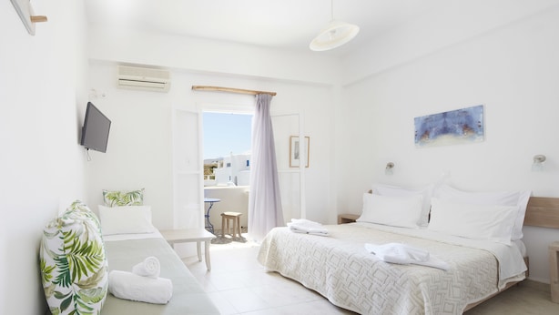 A hotel room with a bed, a bedside table, a TV, and a view of the outside. - Londos (Paros)