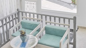 A balcony with a white railing, two teal cushioned chairs, a small round table with a vase, and a view of a street and greenery outside. - Londos (Paros)