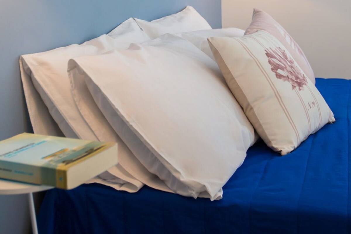 Cama bem arrumada com lençóis brancos e espalha azul, travesseiro com desenho floral e um livro sobre uma mesinha ao lado da cama.