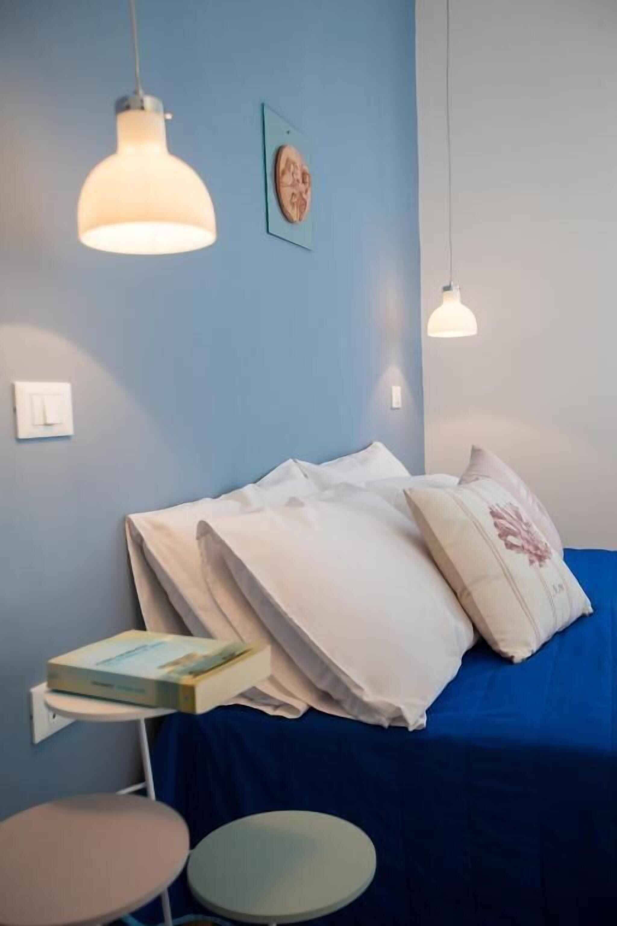 A bedroom with a blue bed, white pillows, a small table with a book, and two hanging lights.