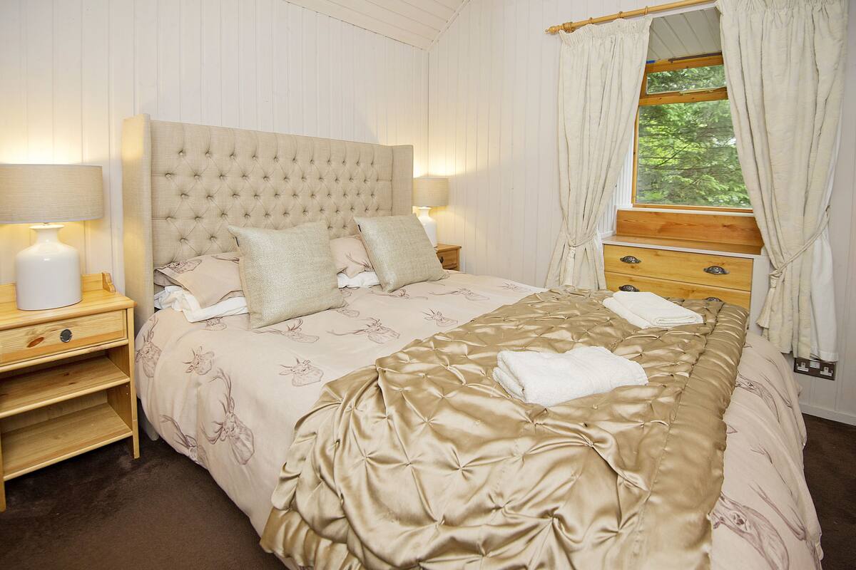 A neatly made bed with a headboard, two bedside tables with lamps, a wooden dresser, and a window with curtains.