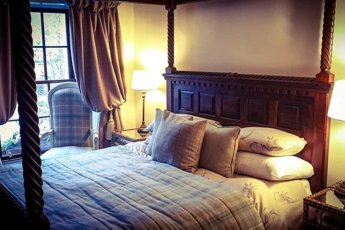 A four-poster bed with a headboard, two bedside lamps, a chair, and a window with curtains.