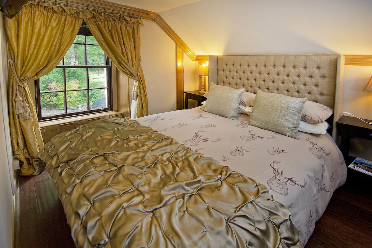 A bedroom with a large bed, a window with curtains, a nightstand with a lamp, and a headboard with a tufted design.