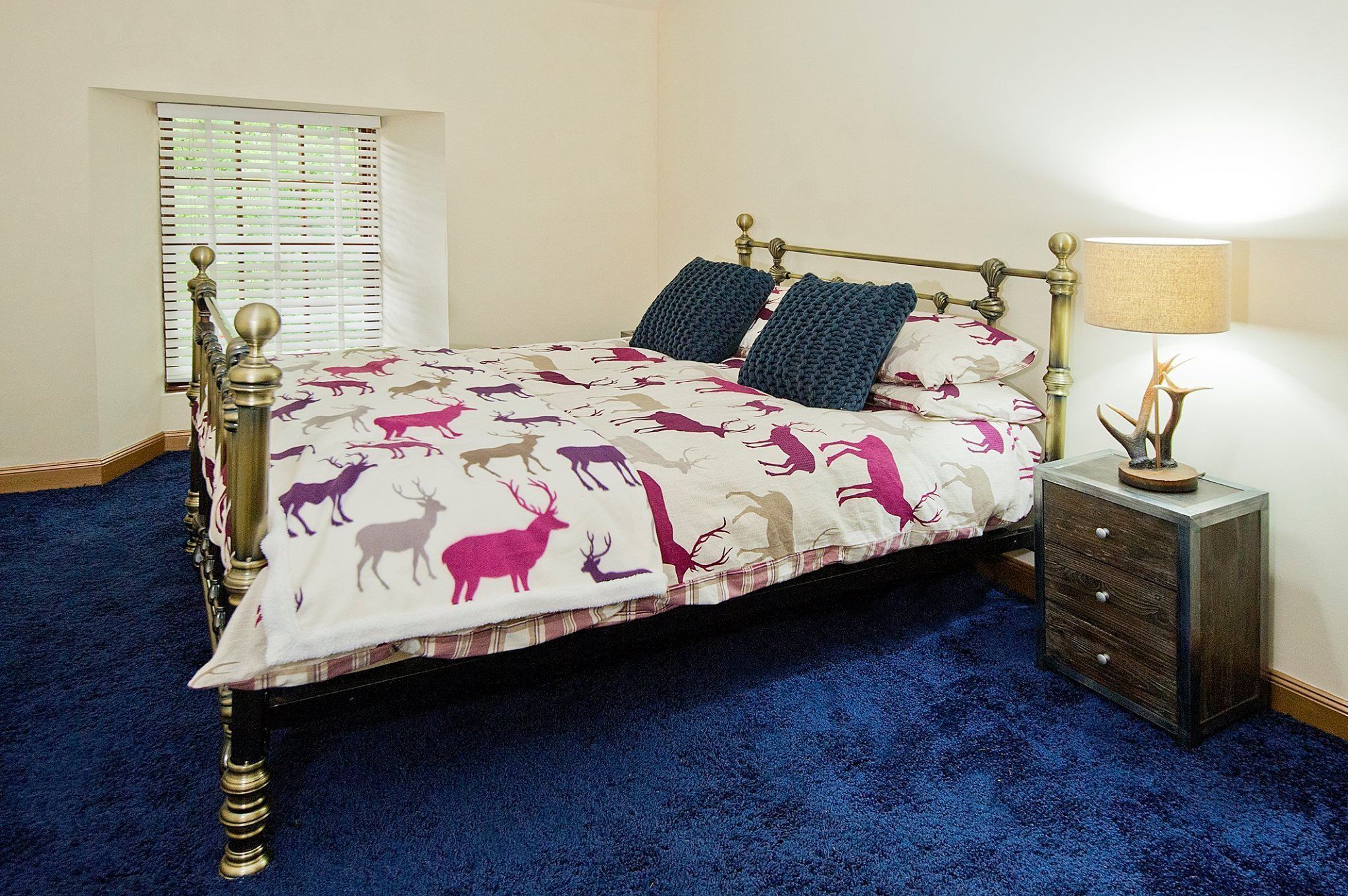 A bed with a patterned bedspread, a bedside table with a lamp, and a window with blinds.