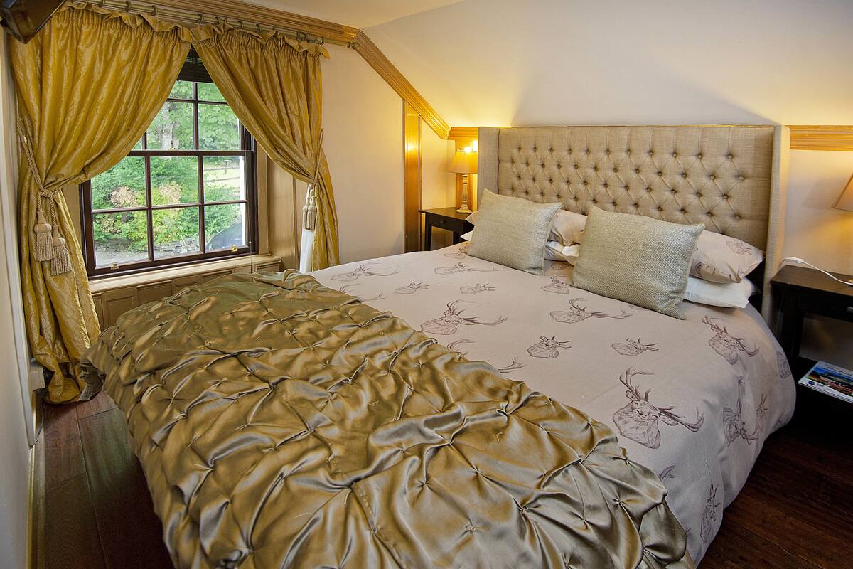 A bedroom with a large bed, a window with curtains, a nightstand with a lamp, and a headboard.