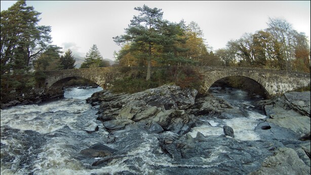 Un puente de piedra sobre un río embravecido, con árboles en los alrededores y un cielo nublado.
