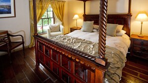 Una cama con dosel sobre una estructura de madera, dos mesitas de noche con lámparas, una silla y un ventanal con cortinas.