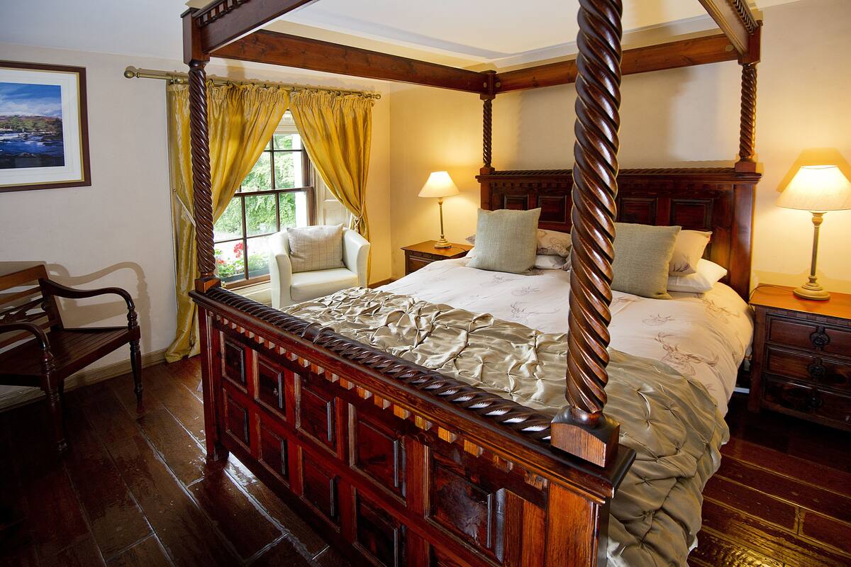A four-poster bed with a wooden frame and a canopy, two bedside tables with lamps, a chair, and a window with curtains.