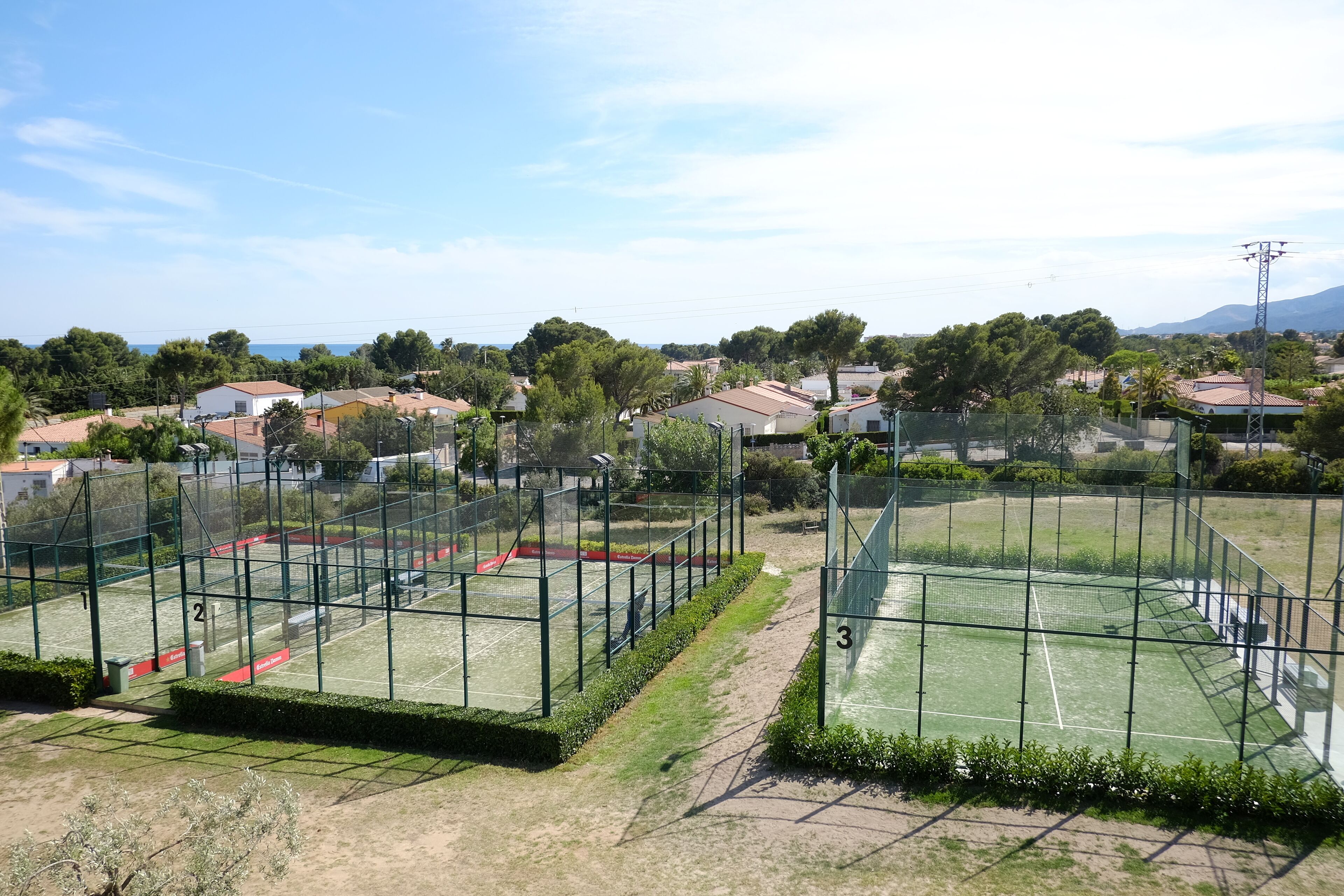 Una cancha de tenis con tres áreas delimitadas, rodeada por una valla, con casas y árboles al fondo.