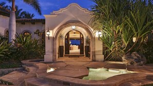 Entrada de hotel con un arco, un camino de piedra que lleva a una piscina y palmeras.