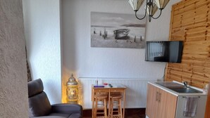 A compact living space with a sofa, a small wooden table, a lamp, a kitchen area with a sink, and a TV mounted on the wall. - The Anchorage (Amlwch)