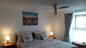 A bedroom with a bed, two bedside lamps, a ceiling fan, a window with curtains, and a painting on the wall. - The Anchorage (Amlwch)