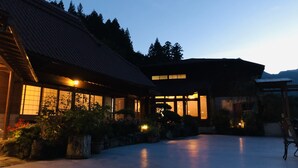 Een traditioneel Japans gebouw met pannendak en grote ramen, omgeven door een binnentuin met stenen lantaarns en potplanten.