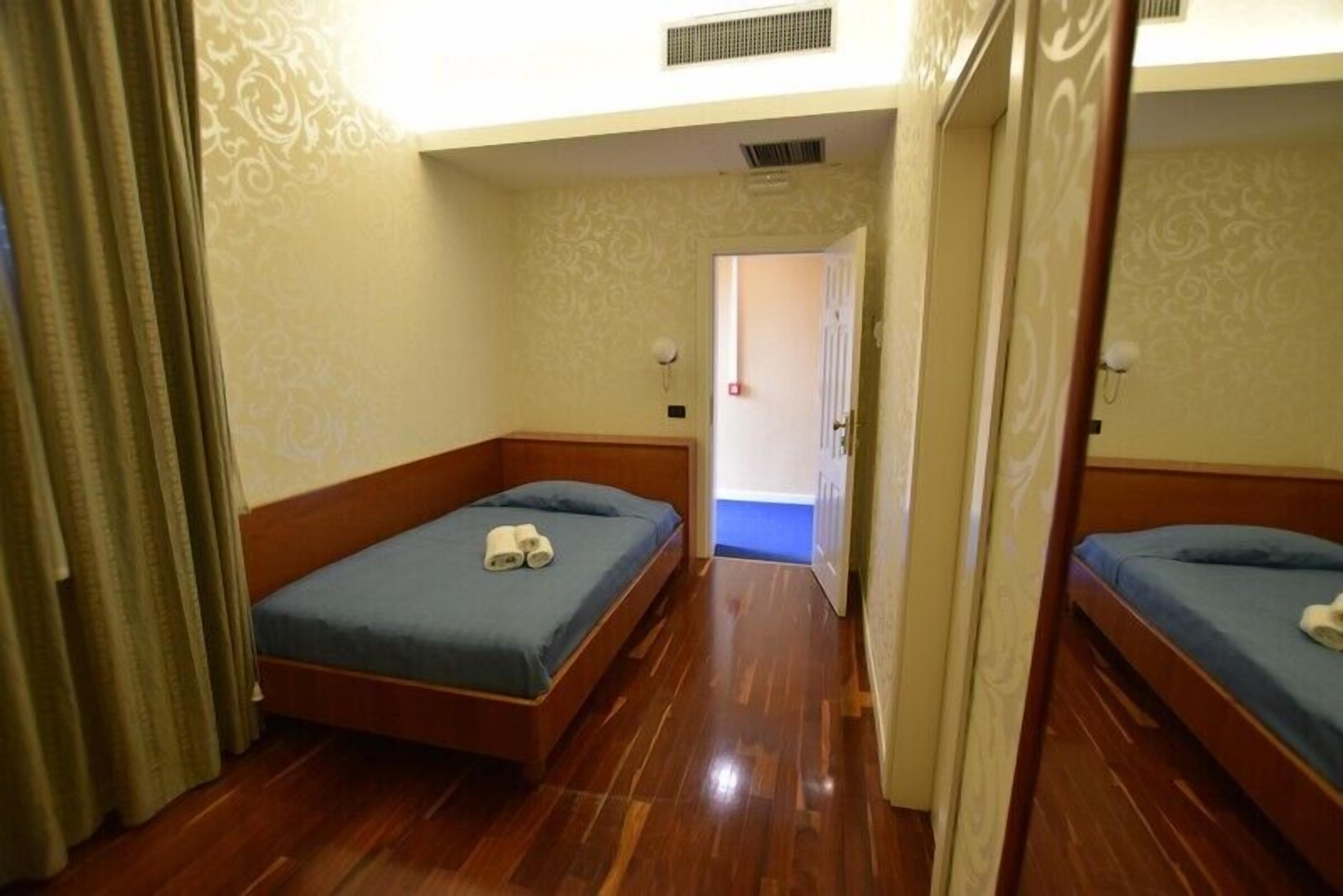 A hotel room with two single beds, wooden flooring, patterned wallpaper, and a door leading to another room.