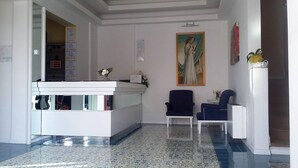 Zona de recepción con un mostrador blanco, dos sillas azules, un cuadro en la pared y piso de baldosas.