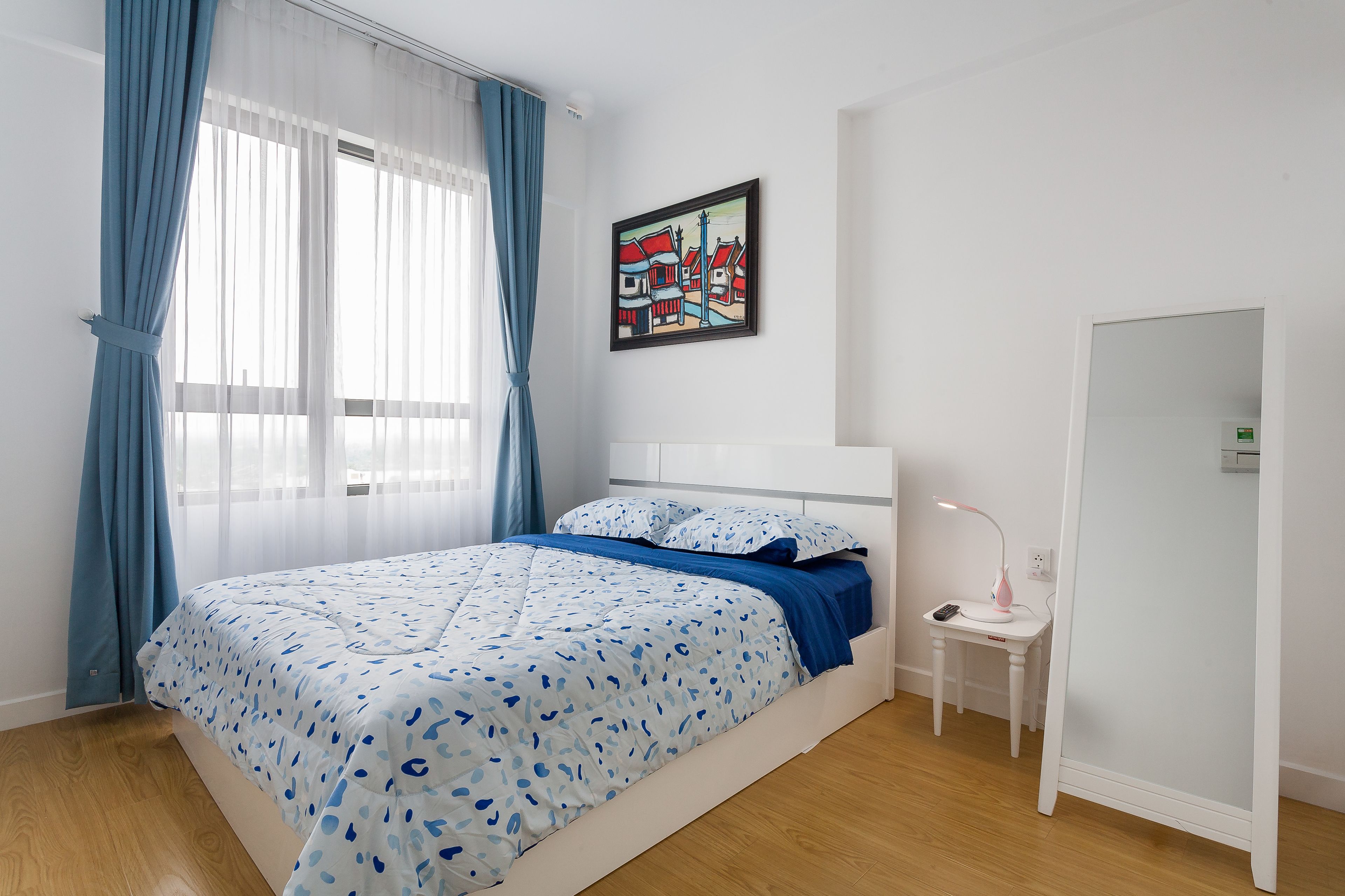 Um quarto com cama, mesa de cabeceira, escrivaninha, cadeira, quadro e porta.