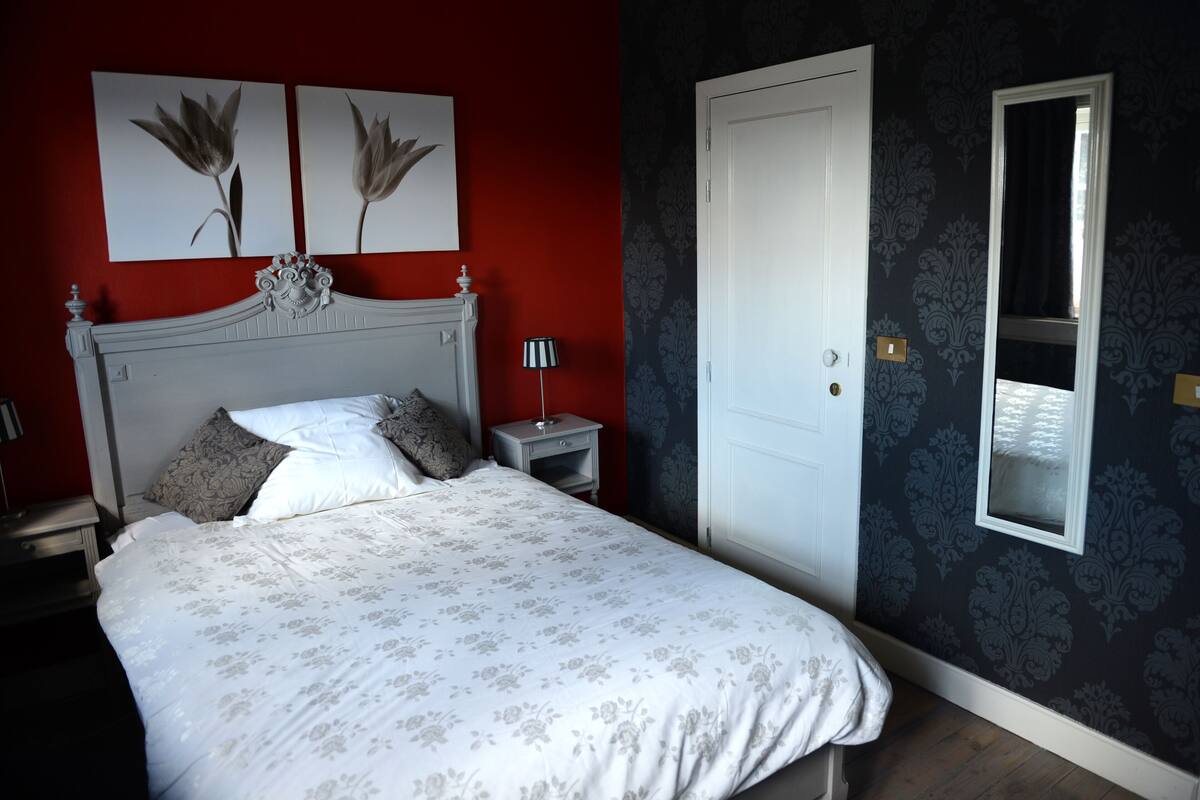 A bedroom with a bed, a nightstand, a lamp, a door, and a mirror.