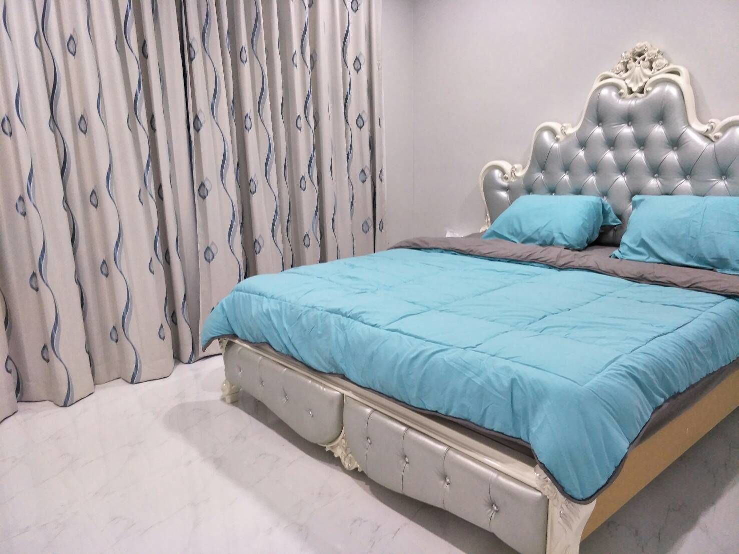Un dormitorio con cabecera capitonada, colcha azul y cortina estampada.