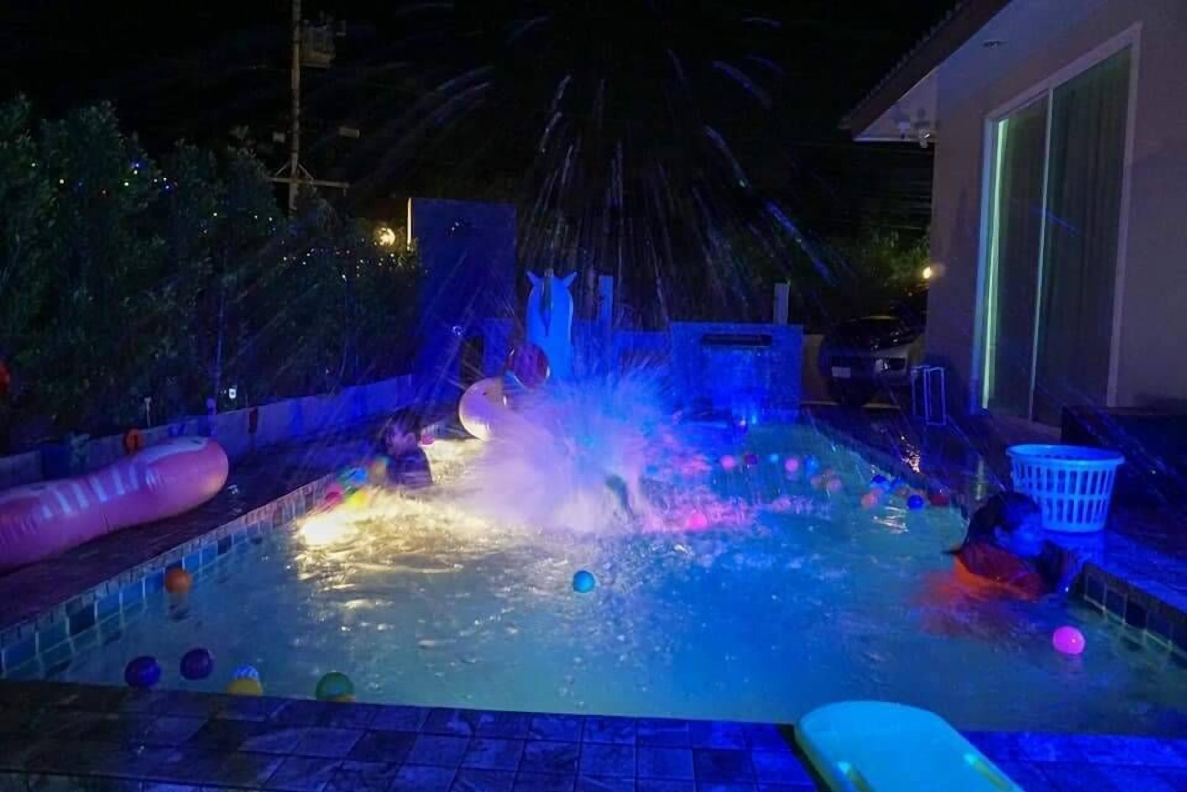 A swimming pool with water jets and colorful balls, surrounded by a fence and trees.