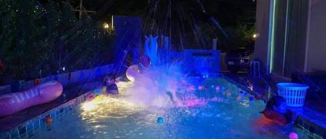 A swimming pool with water jets and colorful balls, surrounded by a fence and trees.