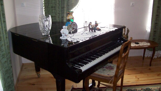 A grand piano with decorative items on its stand, a wooden chair, and a small table with a doily. - Sweet Home (Kazbegi)