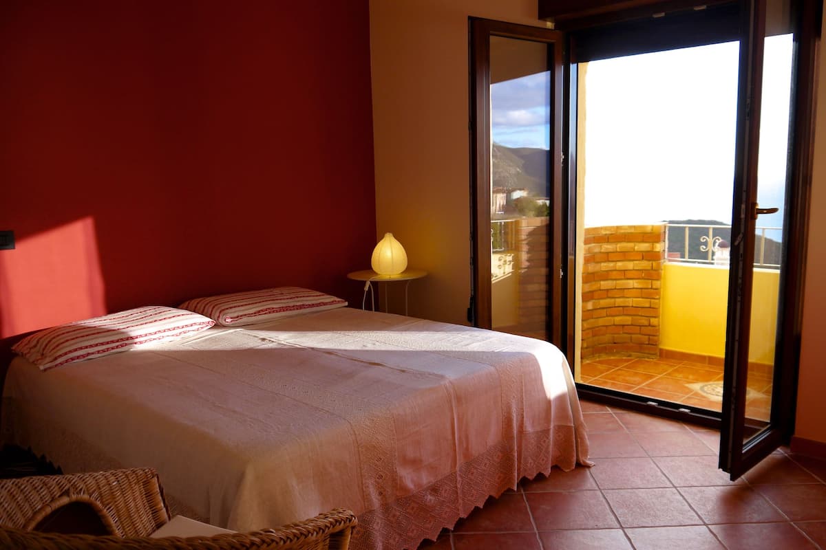 A bedroom with a bed, a nightstand, a lamp, and a view of the outside balcony.