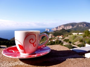 Una taza de cerámica blanca con diseños rojos sobre un plato, colocada sobre una roca con vistas panorámicas a montañas y una masa de agua al fondo.