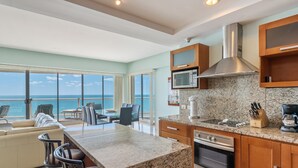 A modern kitchen with granite countertops, stainless steel appliances, and wooden cabinets. It features a large island with bar stools, a dining area with a view of the ocean, and a spacious living area with a sofa and chairs. - Luna Blanca 0801-2BR by Kivoya (Puerto Peñasco)