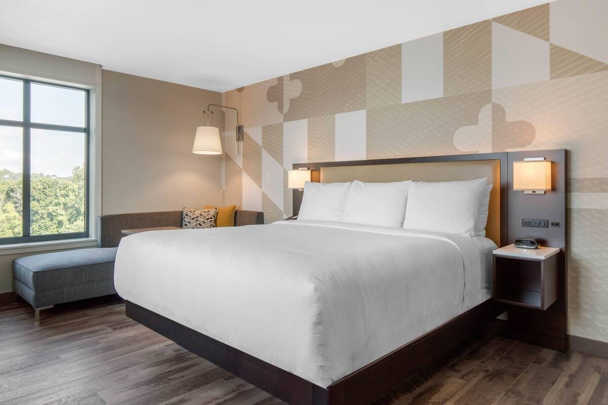 A modern hotel room with a large bed, bedside lamps, a small seating area, and a window with a view of greenery.
