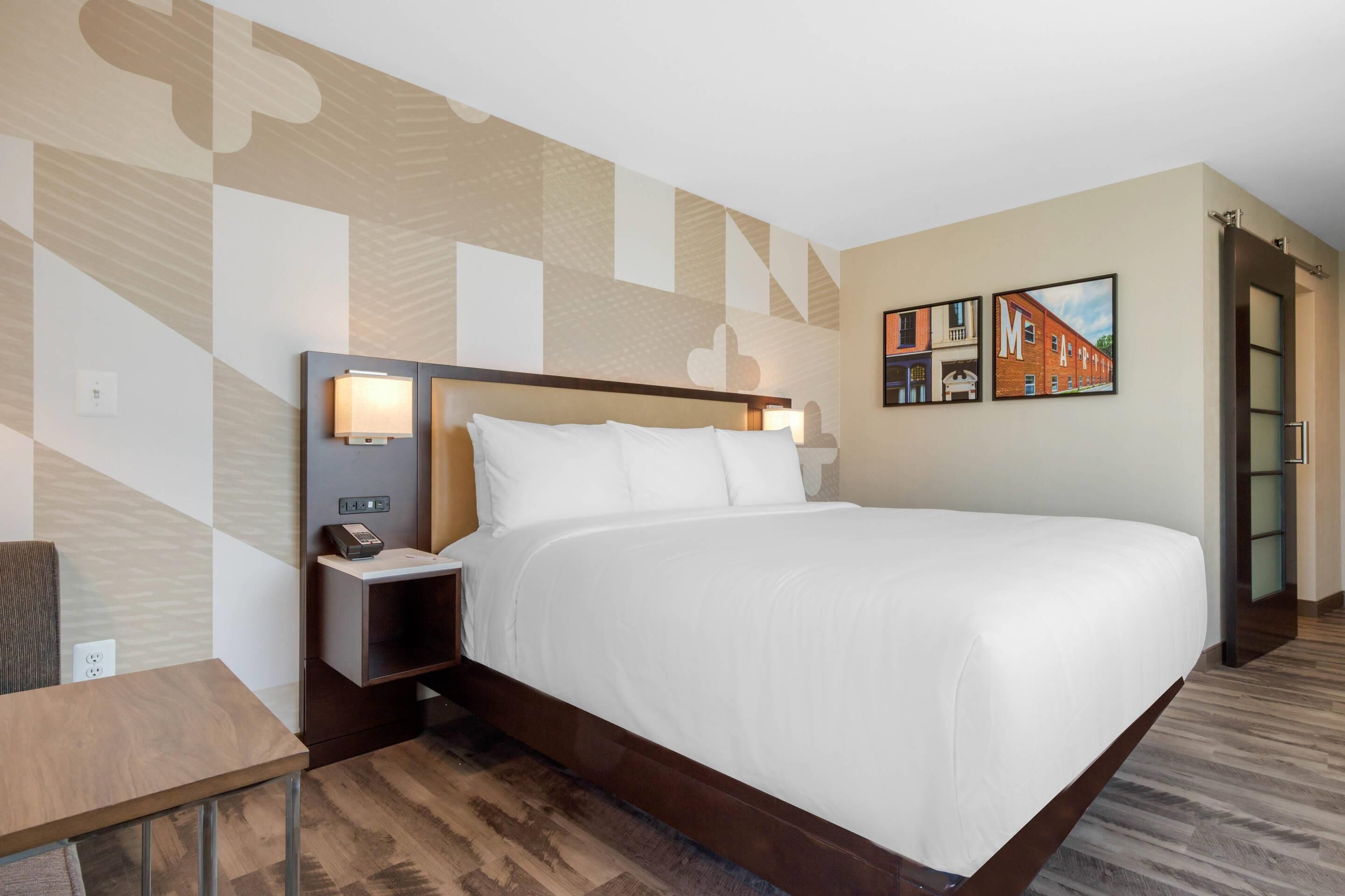 A hotel room with a large bed, a bedside table with a lamp, and a wall with geometric patterns and framed pictures.