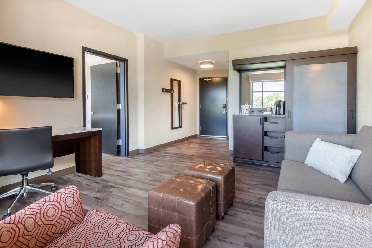 A modern living room with a flat-screen TV, a desk, a sofa, and a kitchen area in the background.