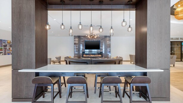 A modern kitchen with a central island, bar stools, hanging pendant lights, and a built-in TV. - Cambria Hotel College Park (College Park)
