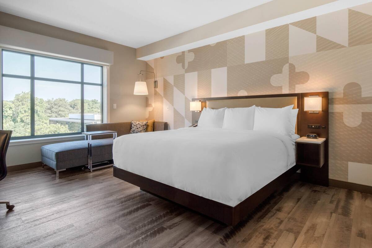 A modern hotel room with a large bed, bedside lamps, a small seating area, and a window offering a view of the outdoors.