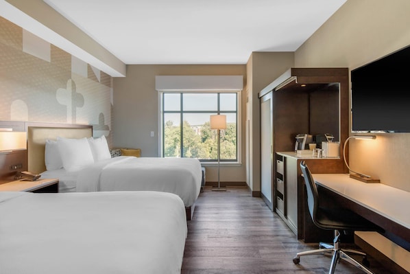 Habitación de hotel con dos camas, un escritorio, una silla, un televisor y ventana con vistas a árboles.