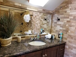 Een badkamer met een granieten aanrechtblad, een wastafel, een grote spiegel, een potplant en toiletartikelen op het aanrecht.
