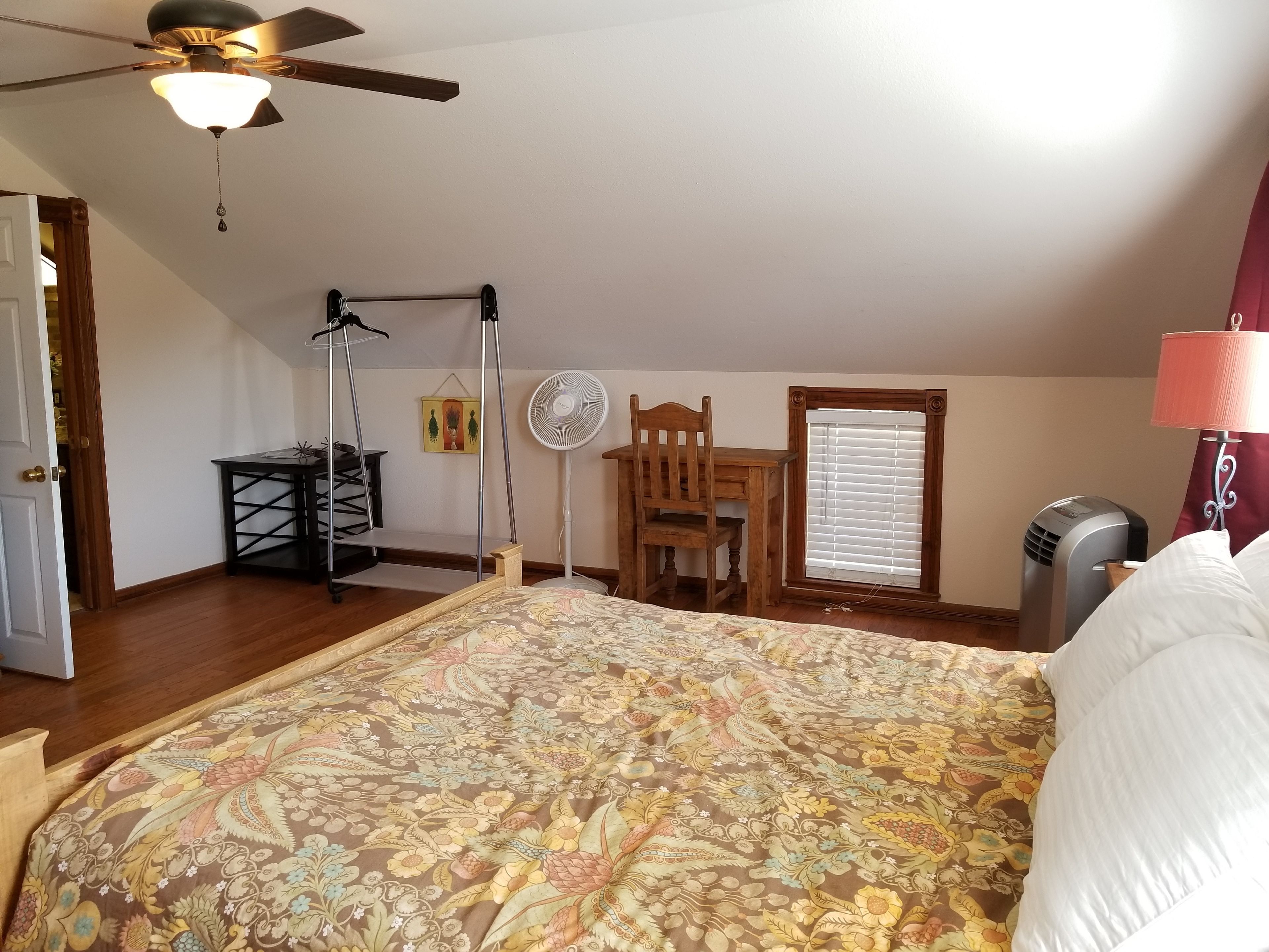 A bedroom with a bed, a ceiling fan, a standing fan, a wooden chair, a table, and a window with blinds.