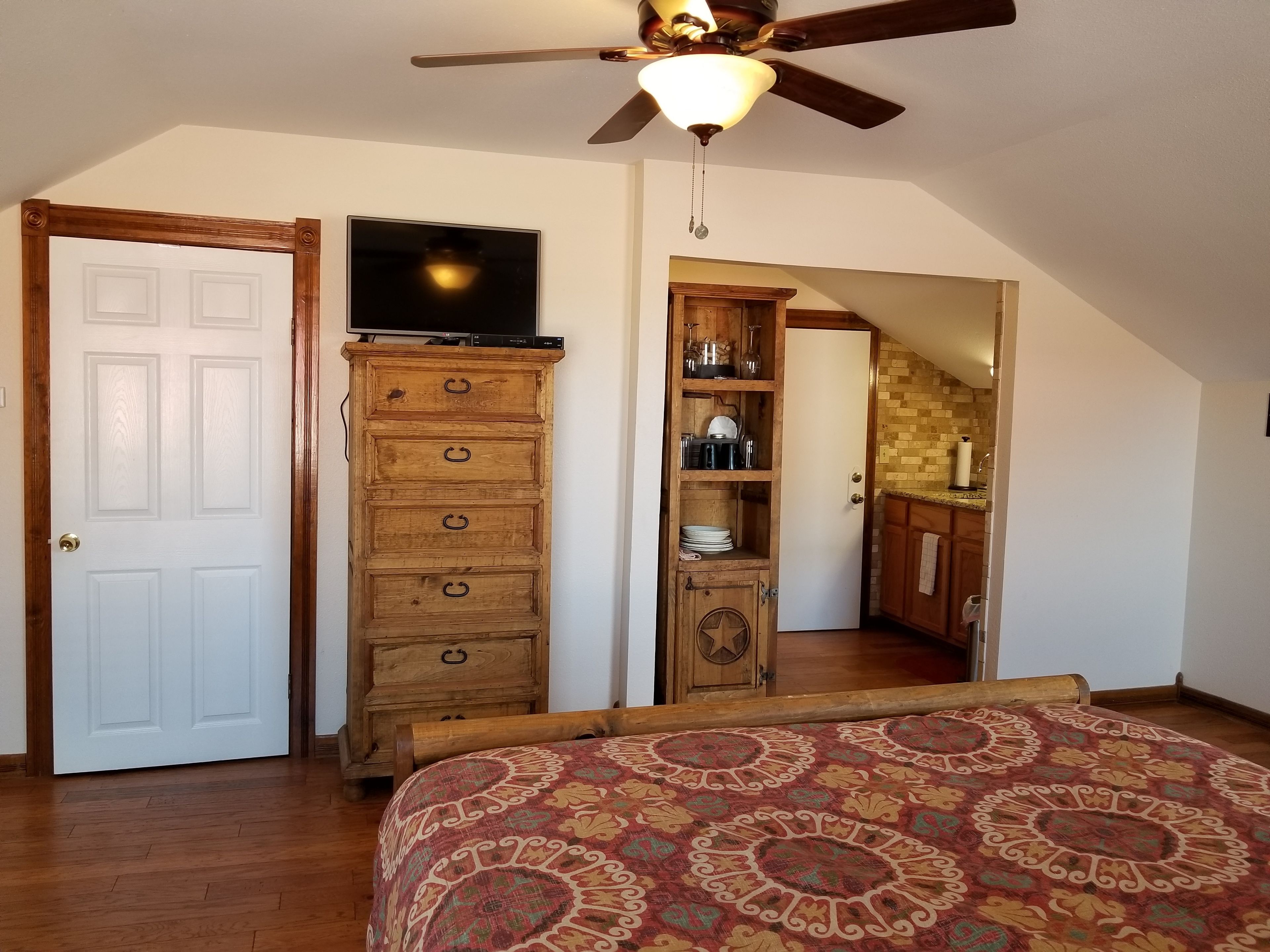 A bedroom with a bed, a wooden dresser, a television on top of the dresser, a ceiling fan, and a door leading to a bathroom.
