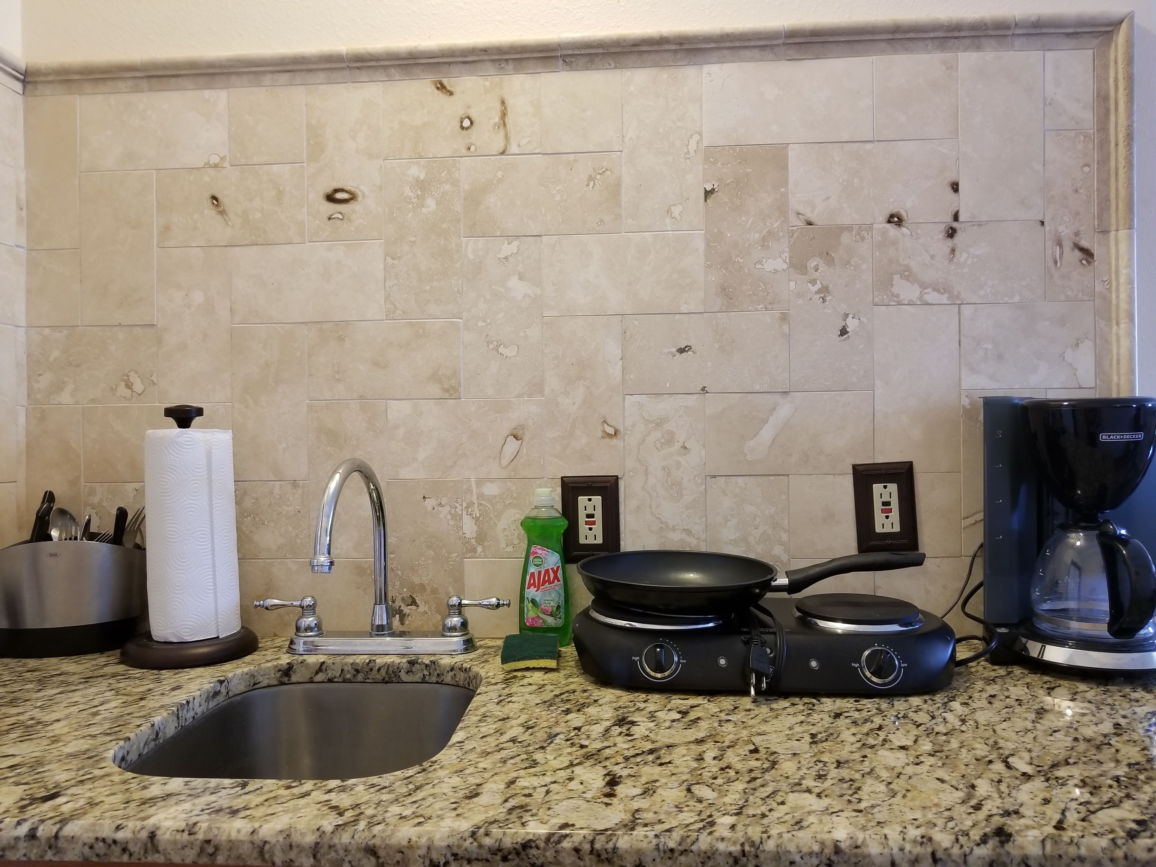 Una cocina con encimera de granito, pileta de acero inoxidable, cafetera, sartén y botella de detergente.