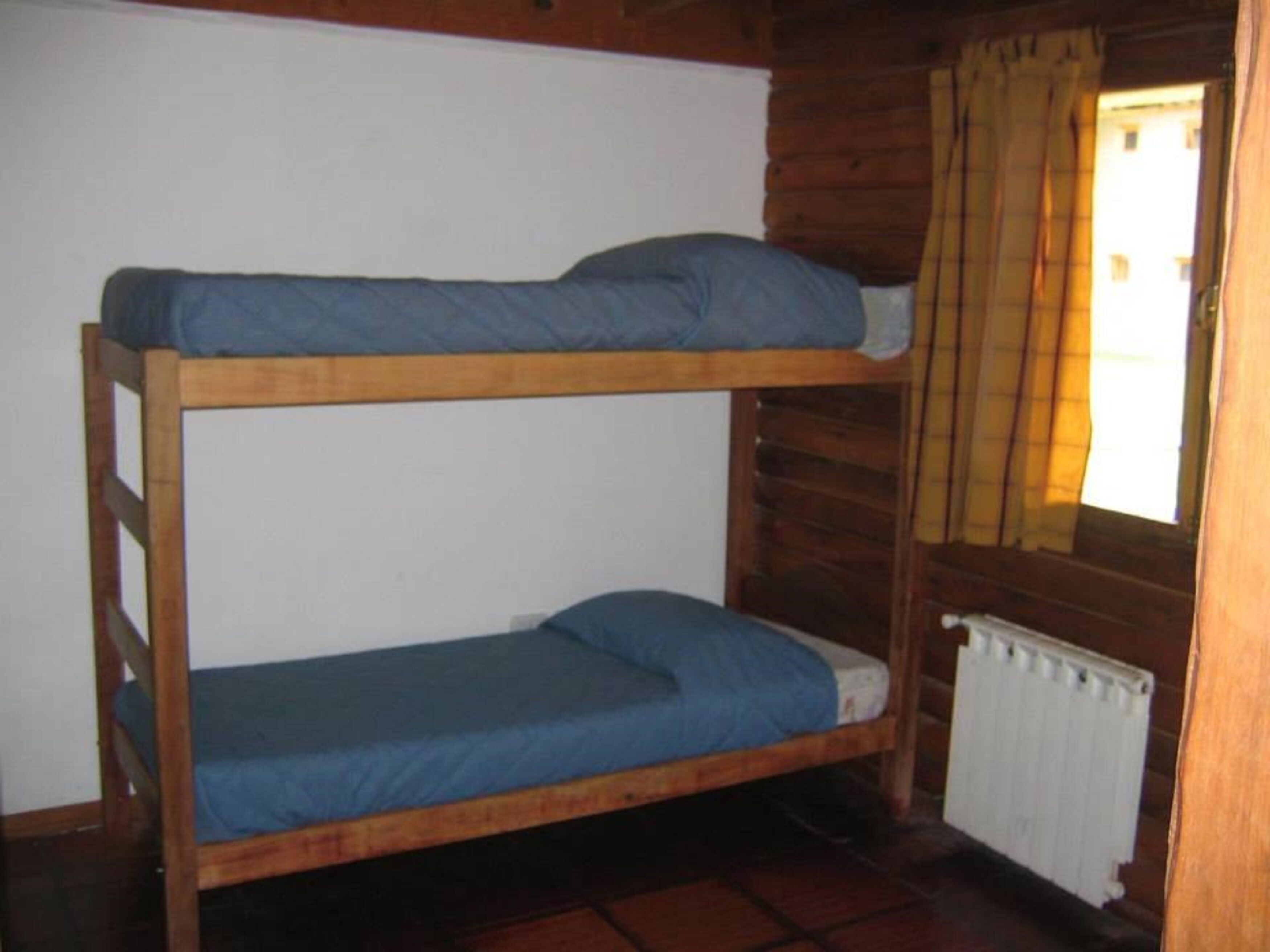 A bunk bed with blue bedding in a cabin-style room with wooden walls and a window with yellow curtains.