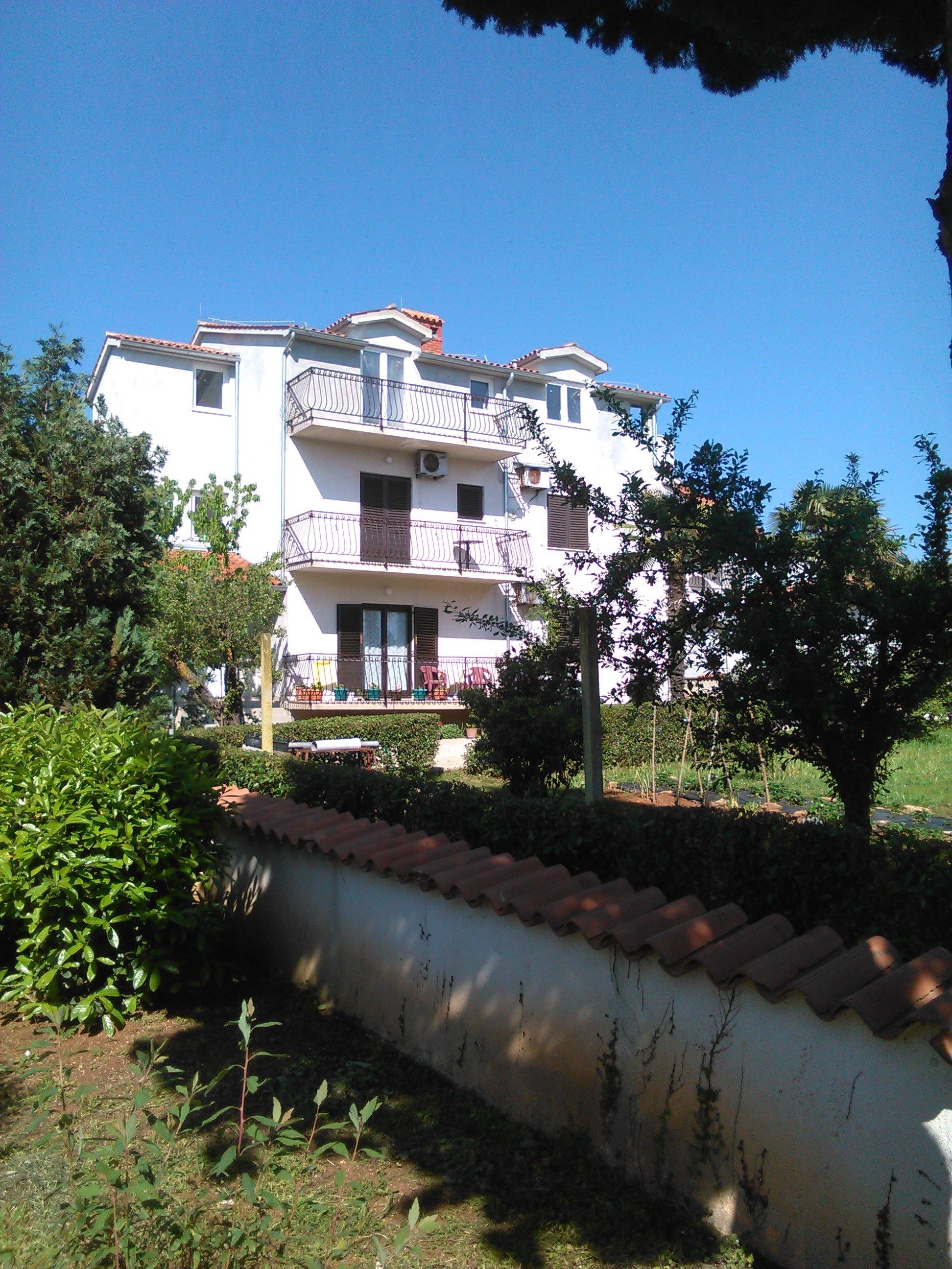 Un edificio de varios pisos con balcones, rodeado de vegetación y un muro bajo.