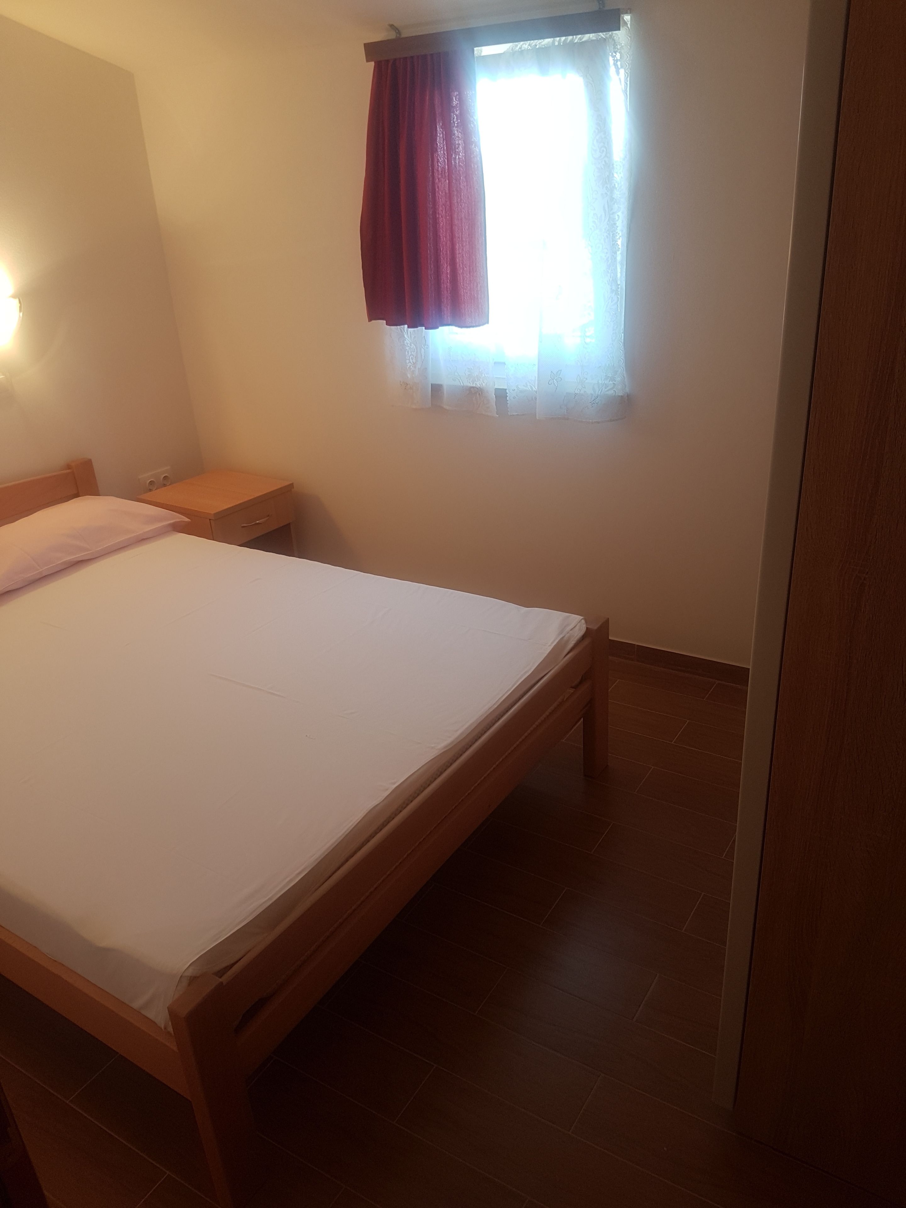 Quarto com uma cama de solteiro, mesa de cabeceira, janela com cortina e tomada na parede.