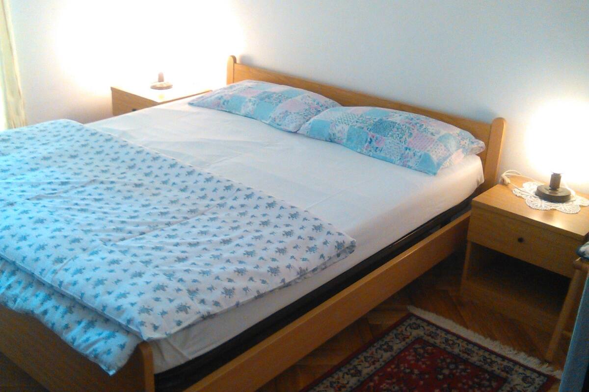 Un lit simple avec une couvre-lit et des oreillers à motifs, une table de chevet en bois avec une lampe, et un tapis rouge et bleu à motifs sur un plancher en bois.