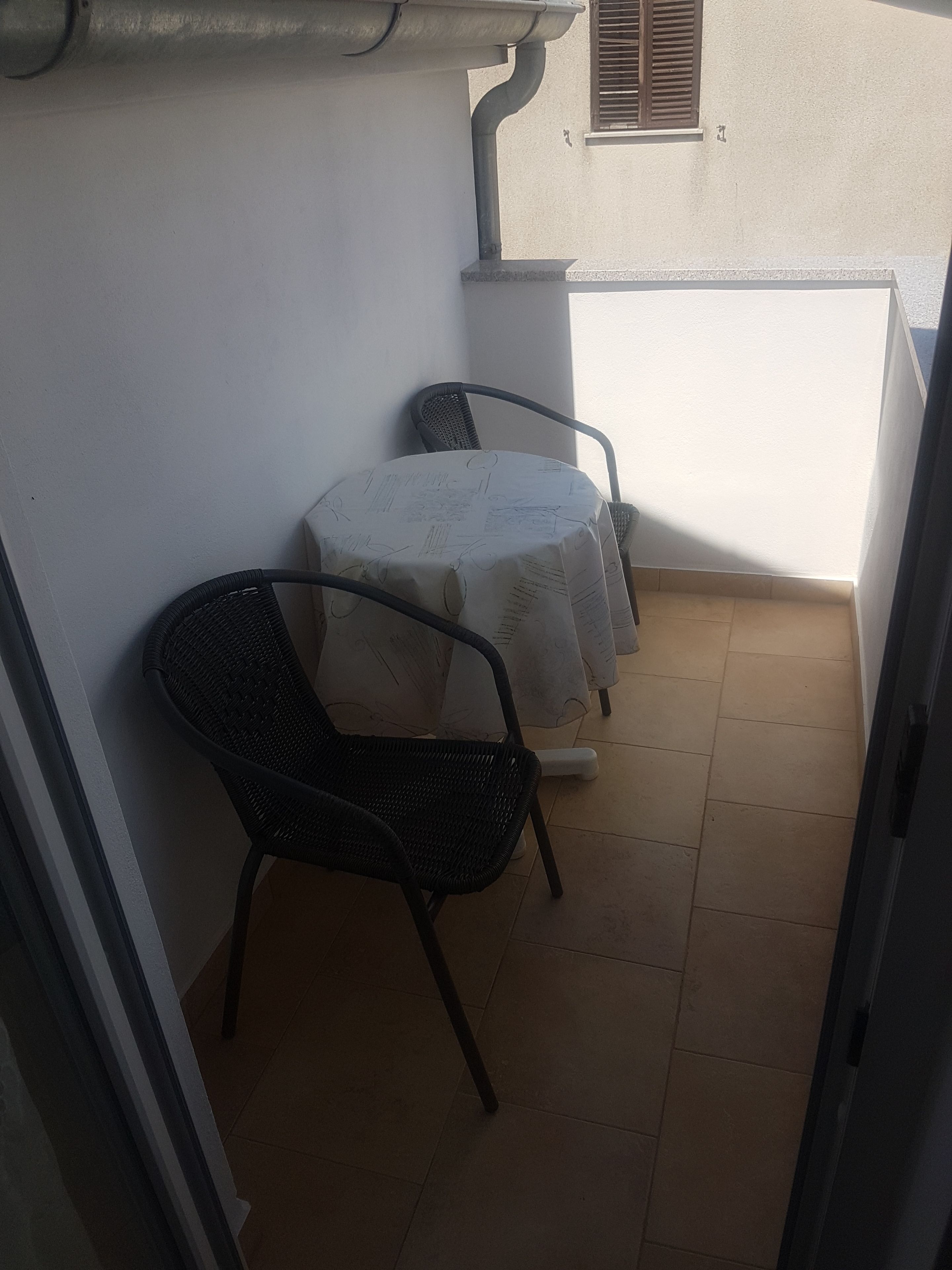 A small balcony with a table and two chairs, a tiled floor, and a white wall with a window.