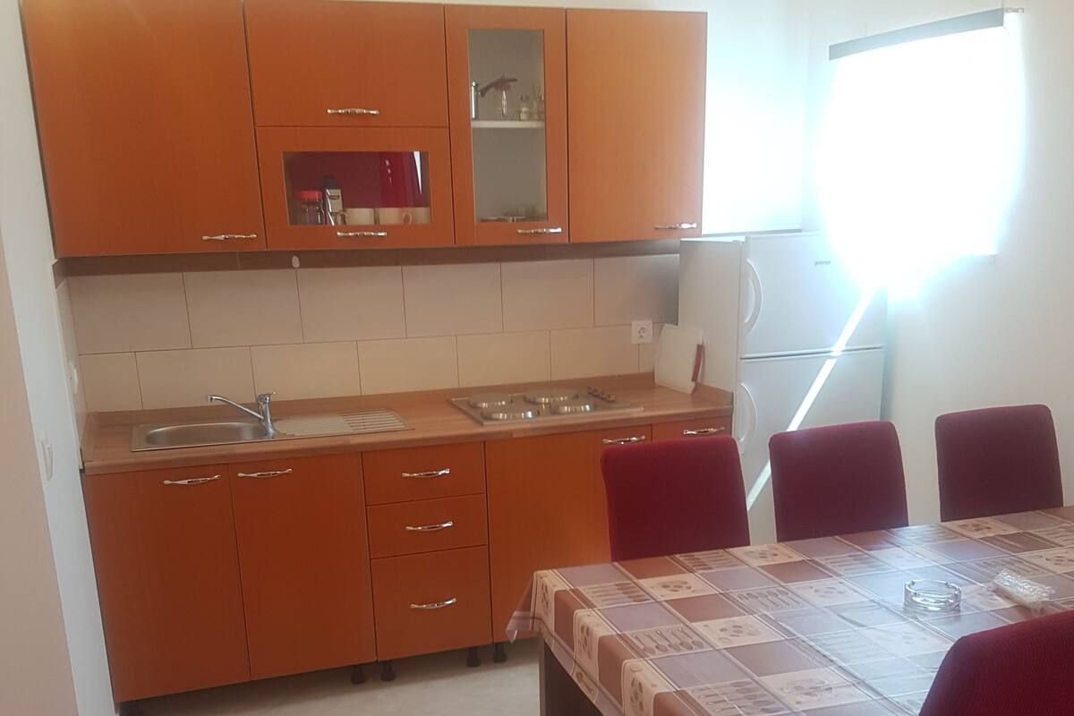 Une cuisine moderne avec des armoires orange, un évier en acier inoxydable et un four encastré. On y trouve une table à manger recouverte d’une nappe à motifs et entourée de chaises rouges.