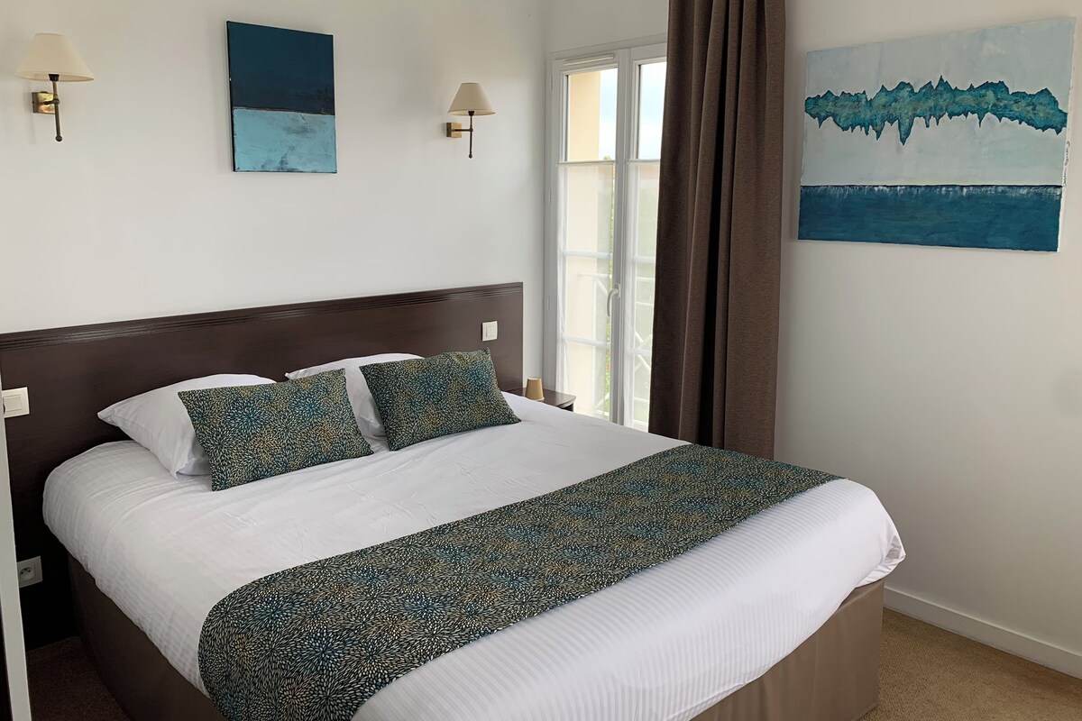 A hotel room with a bed, two wall-mounted lamps, a window with curtains, and two paintings on the wall.