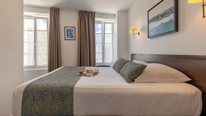 Habitación de hotel bien iluminada con una cama grande, una mesita de noche, una ventana con cortinas, una lámpara de pared y un cuadro enmarcado.