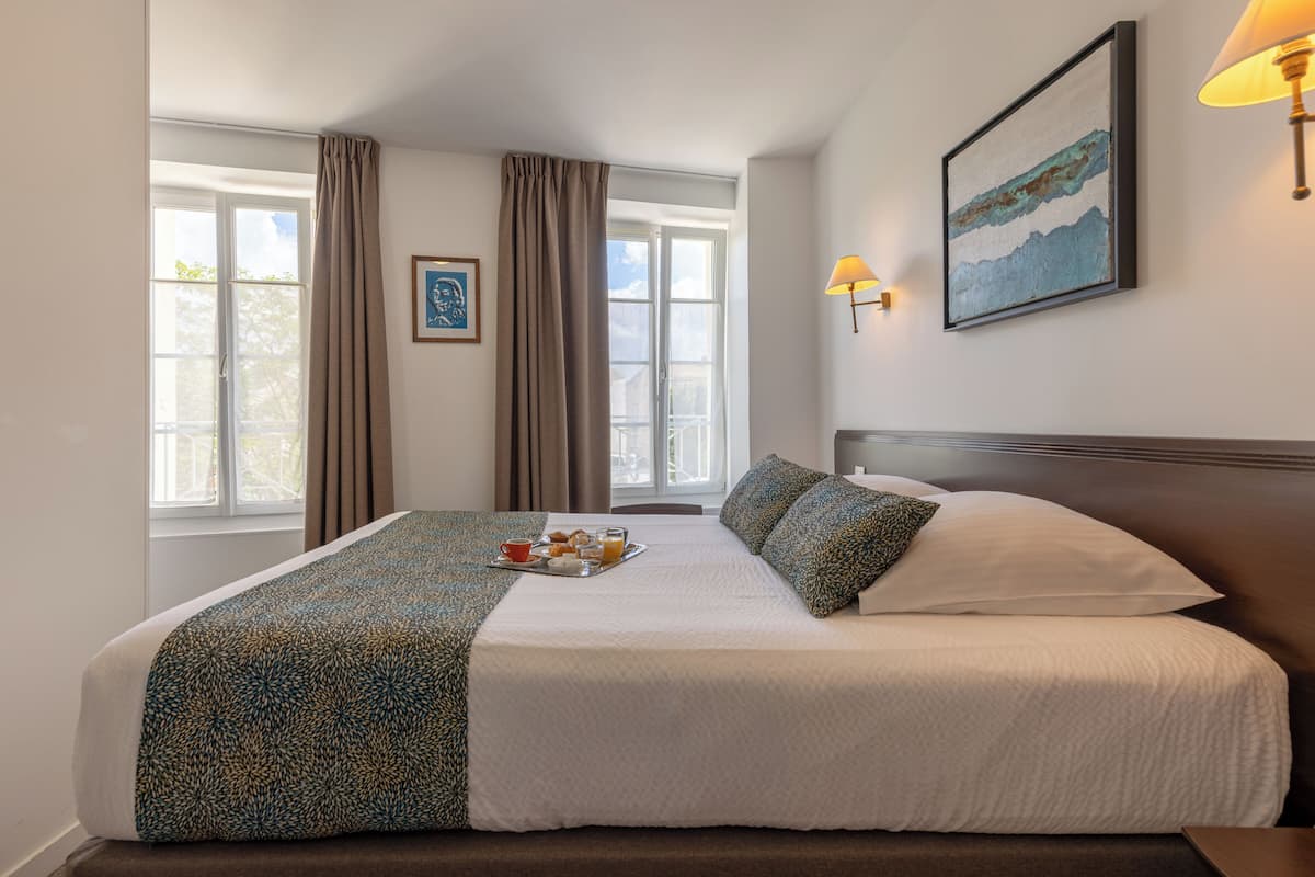 A well-lit hotel room with a large bed, a bedside table, a window with curtains, a wall-mounted lamp, and a framed picture.