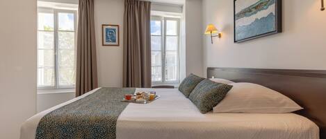 A well-lit hotel room with a large bed, a bedside table, a window with curtains, a wall-mounted lamp, and a framed picture.