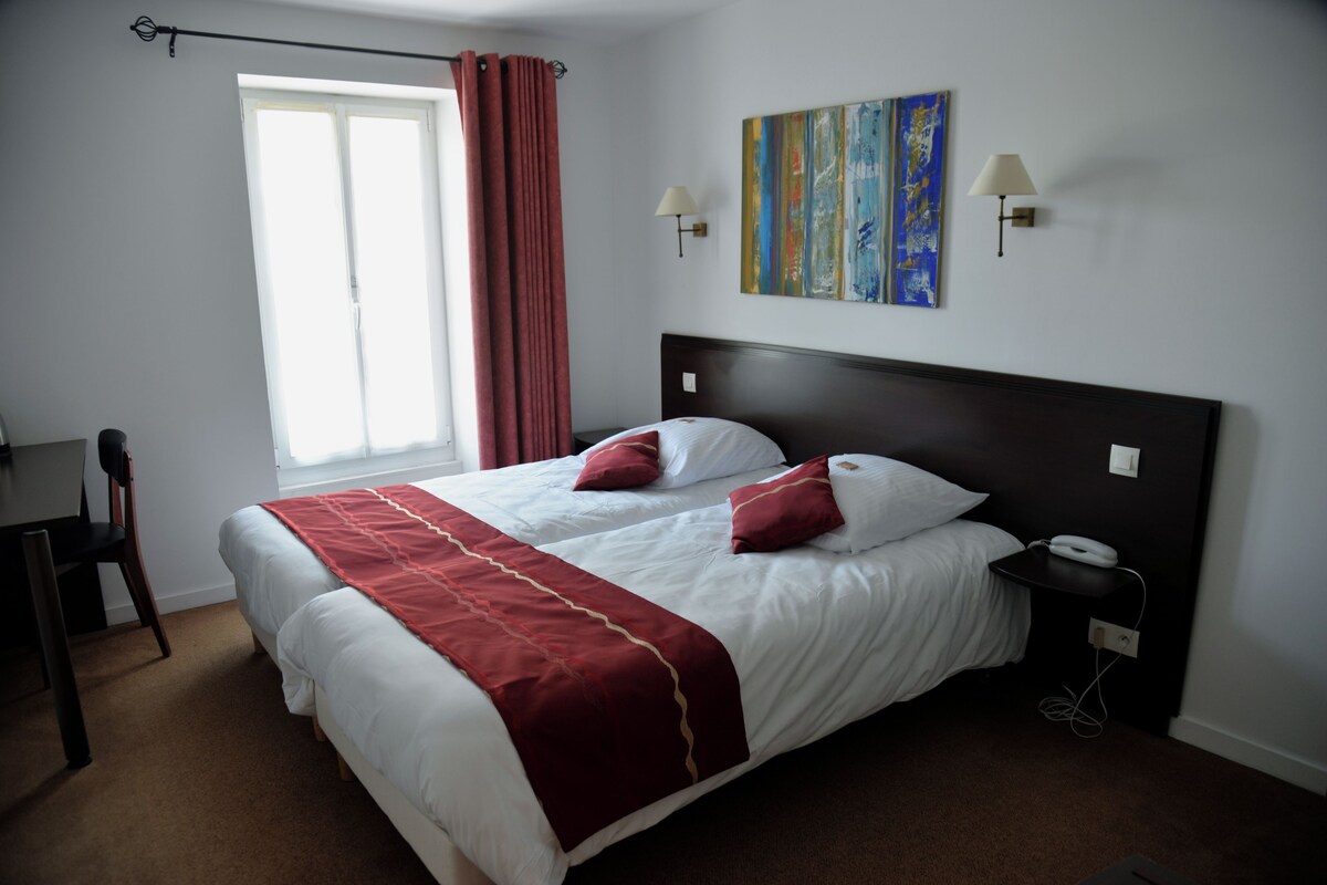 A hotel room with a large bed, two bedside lamps, a nightstand, a desk, and a window with curtains.