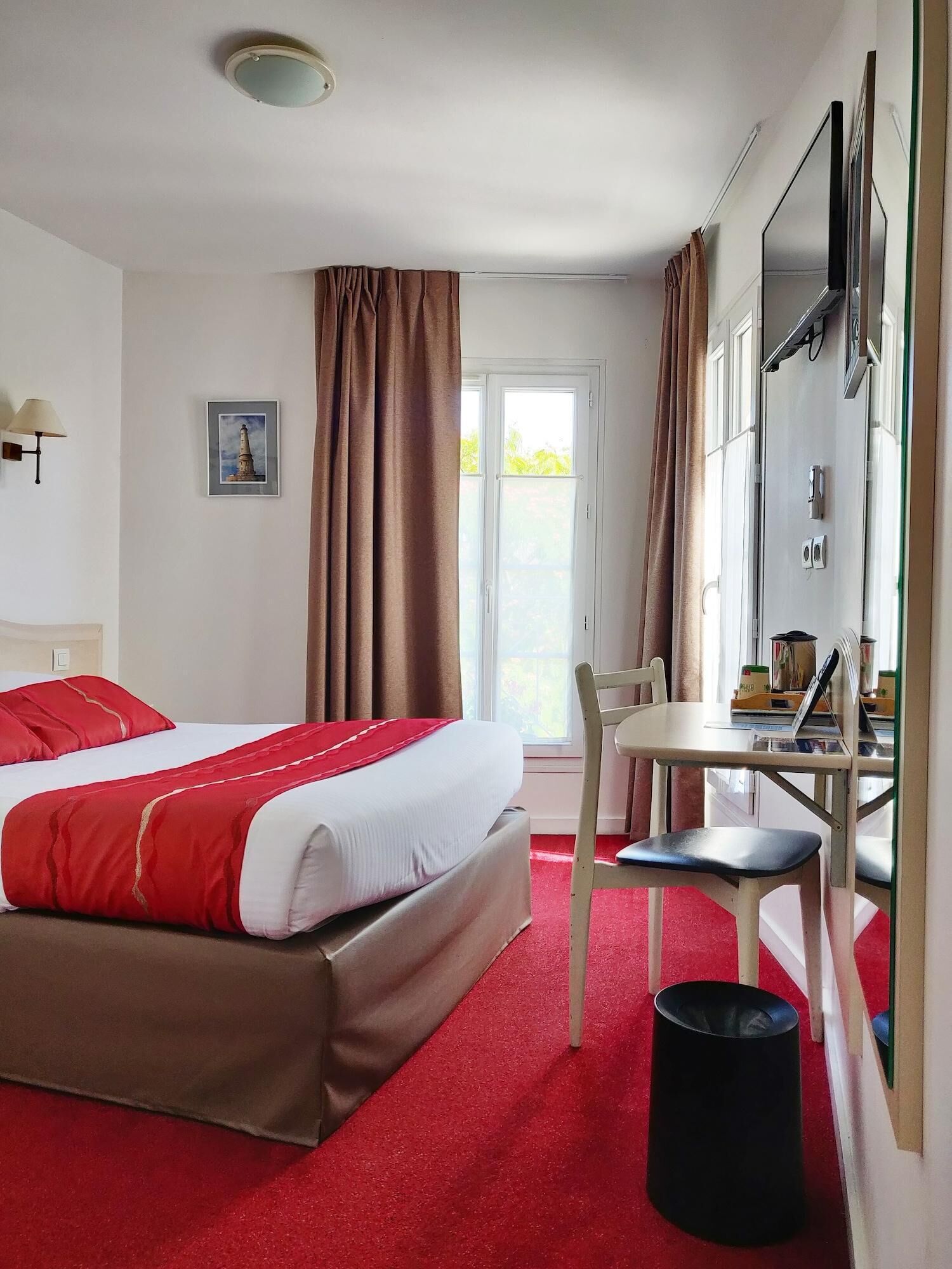 Habitació d'hotel amb un llit, una taula, una cadira, un cubell de brossa i una finestra amb cortines.